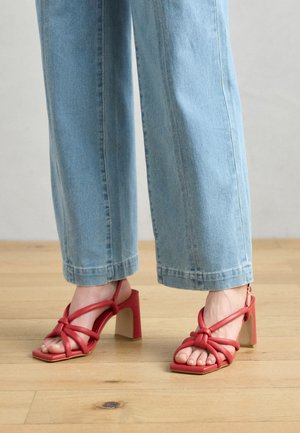 Red heeled sandals with knotted straps and open toe design. Square toe shape and chunky block heel. Worn with light blue wide-leg jeans.