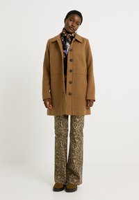 Abrigo de lana marrón con cuello, cierre de botones en la parte delantera y bolsillos laterales; combinado con pantalones de campana con estampado de leopardo y calzado marrón.