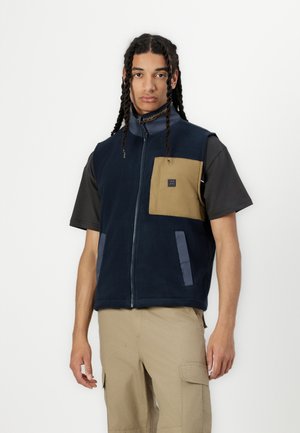 Gilet en polaire bleu marine avec une poche poitrine beige, des accents gris et une fermeture éclair sur toute la longueur à l'avant. Le mannequin porte une chemise noire à manches courtes en dessous.