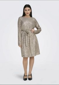 Vestido dourado com lantejoulas, mangas longas, decote redondo e um laço na cintura ajustado. A saia até ao joelho tem uma ligeira evasé. Sapatos de salto alto pretos.