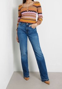 Sweter z wzorem w zygzaki w odcieniach brązu, różu i pomarańczy; połączony z niebieskimi dżinsami w stylu dzwonów i brązowymi szpilkami.