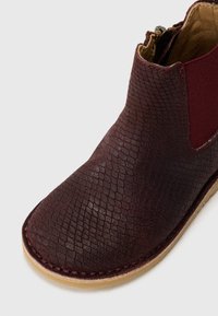 Botín de tobillo burdeos con un upper de cuero texturizado que presenta un patrón similar al de los reptiles, panel lateral elástico y suela de goma beige cosida.