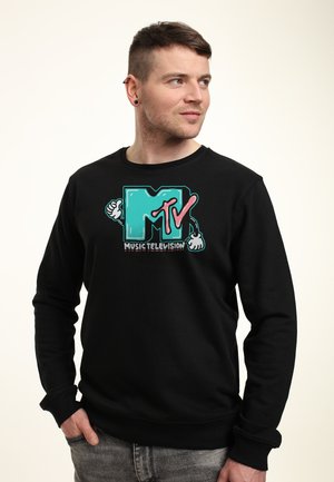 Mann mit kurzen Haaren trägt schwarzen MTV-Sweatshirt mit türkisem Logo, das cartoonartige Arme und den Text "Music Television" zeigt, schaut nach rechts.