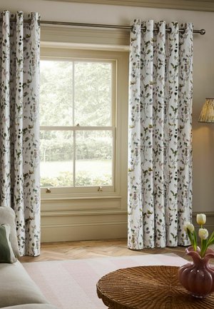Raam met witte gordijnen met bloemmotief, beige muren, houten vloer, ronde rotan tafel met roze vaas en witte bloemen, en beige hoekbank.