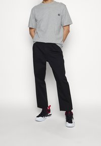 Homme portant un t-shirt gris à manches courtes, un pantalon noir court et des baskets noires, blanches et rouges, debout les mains dans les poches.