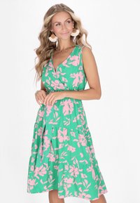 Rochie florală din material verde cu flori roz, fără mâneci, decolteu în V și fustă cu volane. Detalii notabile includ o croială fluidă și o textură moale.