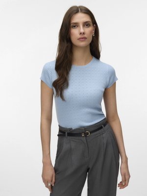 Vrouw met lang bruin haar, gekleed in een lichtblauw kortärmelig gebreid shirt en grijze broek met hoge taille en een zwarte riem, staande tegen een witte achtergrond.