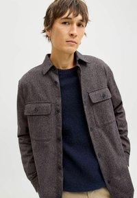 Tumesinine tekstuuriga nööpidega särkkas, millel on kaks rinnataskut, kantud üle mereväe sinise kampsuni. Särgil on mustad nupud ja kraega kaelus.