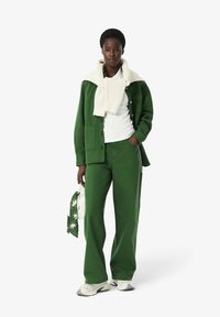 Groene katoenen shirt en broek gecombineerd met een witte gebreide trui om de nek geknoopt. Accessoires omvatten een kleine witte tas en een patroon sjaal.