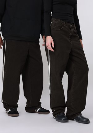 Deux personnes portant des pantalons larges noirs avec des bandes latérales blanches et des hauts noirs, debout côte à côte devant un fond uni.