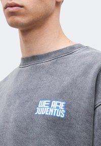 Κοντινό πλάνο ενός ατόμου που φοράει γκρι φούτερ με μπλε κείμενο "WE ARE JUVENTUS" στο στήθος, μπροστά σε απλό φωτεινό φόντο.