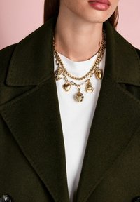 Zelta ķēdes kaklarota ar daudzām piekariņām, tostarp sirdi, kukaiņu un zīdkoku, nēsāta virs baltas krekla un zaļas jakas.