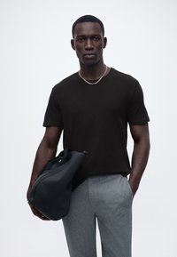 Νέος άνδρας που φοράει μαύρη μπλούζα και γκρι παντελόνι, κρατώντας μαύρη τσάντα στο ένα χέρι, το άλλο χέρι στην τσέπη, στέκεται μπροστά σε απλό λευκό φόντο.