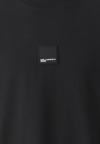 Melnā T-kreklā ar apaļu kakla izgriezumu, izgatavots no mīksta auduma, ar kvadrātveida gumijas logotipa uzlīmi, kurā ir uzraksts "KARL LAGERFELD JEANS" uz krūtīm.
