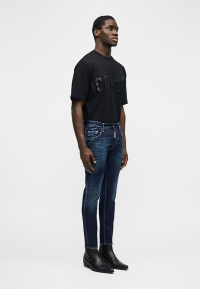 Homme debout portant un t-shirt graphique noir, un jean slim bleu foncé et des bottines en cuir noir, sur un fond gris uni.