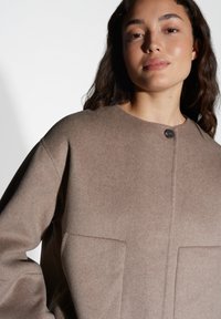Bruine wolmix oversized jas met een ronde halslijn, enkele knoopsluiting en grote voorkruiken. Zachte textuur, minimale naden.