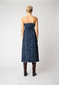 Trägerloses Denim-Kleid mit wirbelnden blauen Mustern, Reißverschluss hinten und Midi-Länge. Kombiniert mit kniehohen braunen Stiefeln mit Absatz.