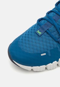 Blauer Sportschuh mit einem Obermaterial aus Mesh, strukturiertem Stoff und einer weißen Gummisohle. Bemerkenswerte Details sind grüne Logoakzente und ein Schnürsystem.