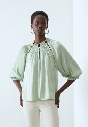 Blusa verde chiaro realizzata in tessuto morbido, con dettagli arricciati sul collo, maniche a sbuffo e accenti a intaglio.