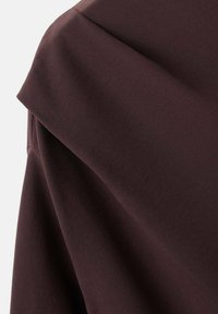 Tela marrón oscura con una textura suave; presenta un diseño holgado y caído con secciones superpuestas, enfatizando su forma fluida.