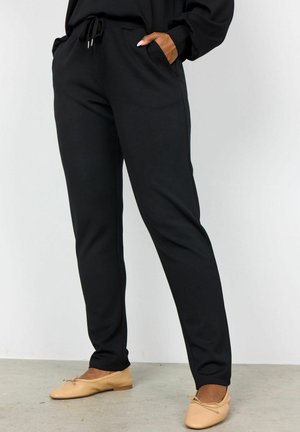 Persoon draagt zwarte broek met trekkoorden, handen in de zakken en beige ballerina's, staand op een grijze vloer tegen een witte muur.