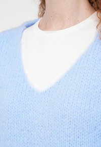 Κοντινό πλάνο ενός ατόμου που φοράει λευκό μπλουζάκι με στρογγυλή λαιμόκοψη κάτω από ένα ανοιχτό μπλε πλεκτό πουλόβερ με V-λαιμό, που δείχνει τον λαιμό και το άνω μέρος του στήθους.