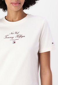 Cremefarvet bomulds t-shirt med korte ærmer. Har maroon tekst "New York Tommy Hilfiger" og lille logo-detalje på ærmet.