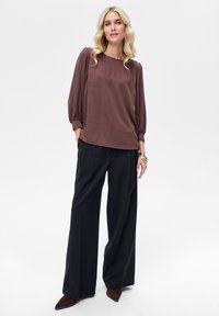 Brązowa satynowa bluzka z rękawami o długości do łokcia typu balloon, zestawiona z czarnymi spodniami o szerokich nogawkach i brązowymi szpilkami z ostrym noskiem, tworząca smukłą sylwetkę.
