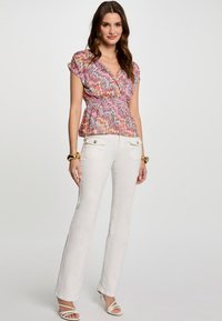 Haut péplum multicolore avec un motif en zigzag, associé à un pantalon blanc évasé et des talons blancs à lanières. Les accessoires comprennent un bracelet en or.