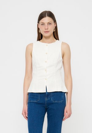 Γυναίκα με ίσια καστανά μαλλιά, που φορά χρυσούς κρίκους στα αυτιά, λευκό αμάνικο πουκάμισο με κουμπιά και μπλε τζιν, που στέκεται μπροστά σε λευκό φόντο.