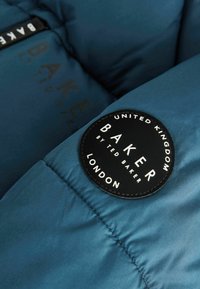 Doudoune en matériau bleu profond, avec un patch logo rond noir et texte blanc, et une fermeture éclair avec une marque sur le ruban.