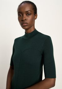 Woman with braided hair wearing a dark green short-sleeve mock-neck top, standing against a plain light background.