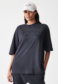 Tamno siva oversized majica s kratkim rukavima s izvezenim logom "NEW ERA" u odgovarajućoj niti, i okruglim izrezom. Glatka pamučna tkanina.