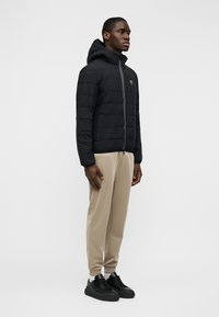 Μαύρο μπουφάν με επένδυση και κουκούλα, με φερμουάρ στο μπροστινό μέρος και ανοιχτόχρωμη ρίγα διακόσμησης· συνδυασμένο με μπεζ παντελόνι στενής γραμμής και μαύρα αθλητικά παπούτσια.