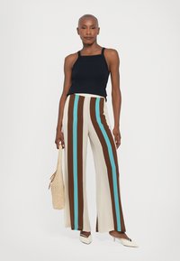 Pantaloni largi de culoare crem cu dungi verticale maro și turcoaz; asortați cu un top negru fără mâneci și pantofi albi cu toc. Poartă o geantă țesută în mână.