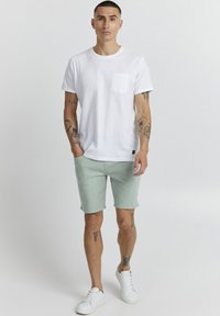 Tricou alb din bumbac cu buzunar la piept, asortat cu pantaloni scurți verzi deschis și adidași albi, având tatuaje pe brațe și picioare.