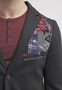 Blazer nero con molteplici tasche applicate in vari tessuti, inclusi plaid e houndstooth, caratterizzato da testo e grafiche stampate. Top bordeaux sottostante.