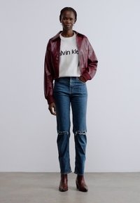 Burgundi nahast pommitaja jope, valge dressipluus musta tekstiga, kõrge vöökohaga sinised teksapüksid rebenenud põlvedega, pruunid pahkluu saapad.