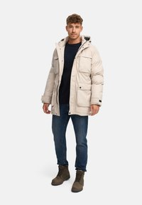 Giacca puffer beige chiaro con cappuccio, multiple tasche frontali e chiusure a scatto, indossata sopra un maglione blu navy, abbinata a jeans blu e stivali marroni.