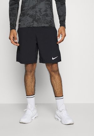 Mężczyzna ubrany w czarne spodenki Nike, białe buty sportowe, białe skarpetki z czarnymi paskami i długim rękawem szarą koszulkę w wzory, stojący przy białej ścianie.