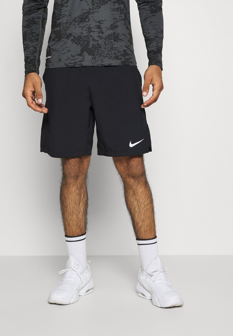 Homme portant un short noir Nike, des chaussures de sport blanches, des chaussettes blanches à rayures noires et une chemise grise à manches longues à motifs, debout contre un mur blanc.
