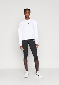 Λευκό φούτερ με μαύρο λογότυπο, σε συνδυασμό με μαύρα κολάν με διακοσμητικά στοιχεία από δίχτυ και λευκές ρίγες, φορεμένα με λευκά αθλητικά παπούτσια.