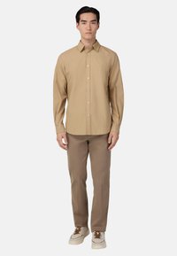 Béžová košile s dlouhým rukávem a límečkem, zapínání na knoflíky, s jemnou strukturou, spárovaná s hnědými kalhotami a bílými teniskami.