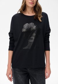 Czarna bluzka z długim rękawem, wykonana z miękkiej tkaniny, z teksturowanym graficznym wzorem przedstawiającym twarz oraz abstrakcyjne kształty w odcieniach szarości.