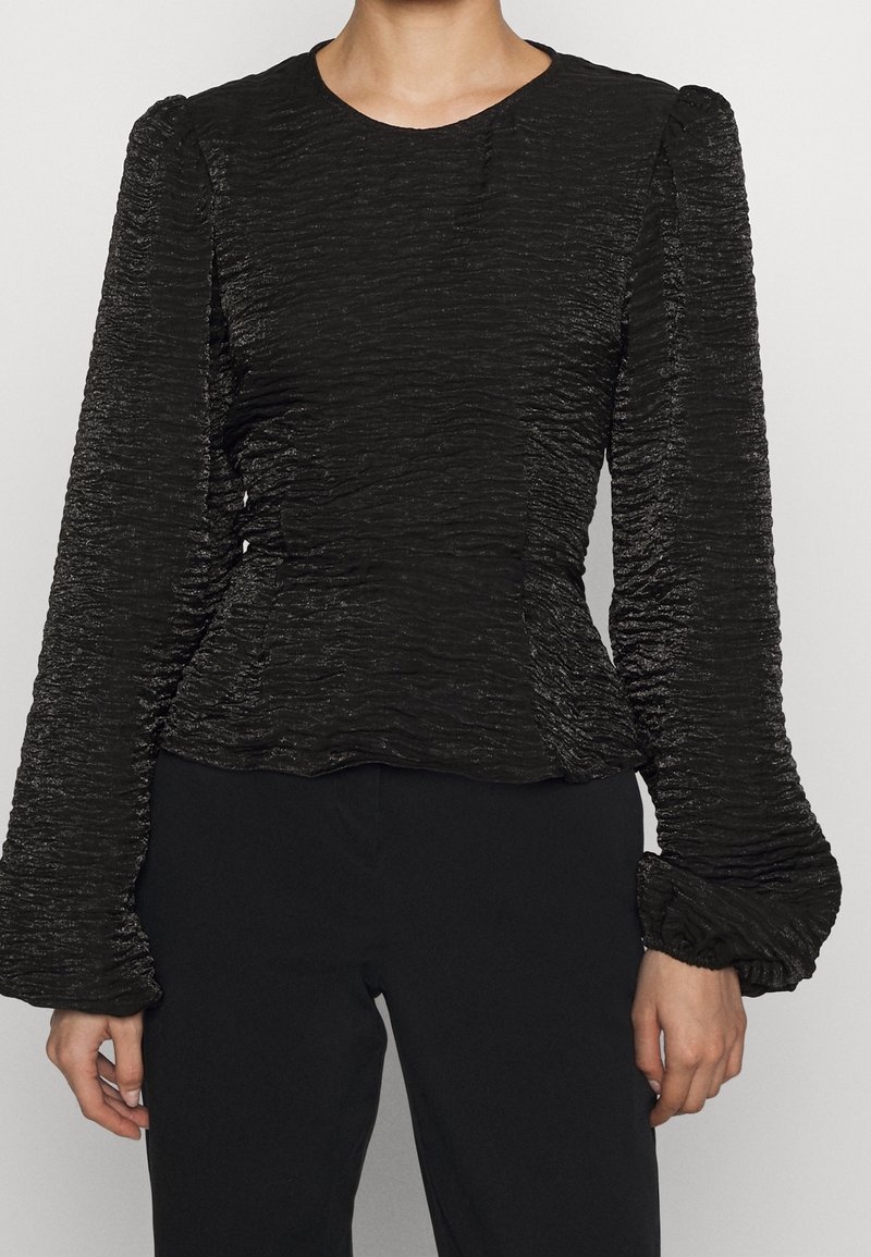 Femme portant un chemisier noir texturé à manches longues bouffantes et taille cintrée, associé à un pantalon noir, sur un fond uni.