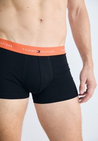 Sorte bomuldsboxershorts med en orange elastisk talje, der har "TOMMY HILFIGER" logo. Glat tekstur, tætsiddende design.