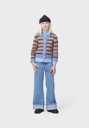 Jeune personne debout portant un bonnet bleu marine, un cardigan rayé boutonné, une chemise bleu clair, un jean large avec revers et des chaussures noires sur fond uni.