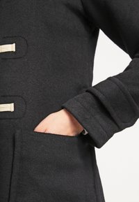Cappotto di lana nero con tasca laterale, texture tonale prominente e dettagli di chiusura a toggles beige sul davanti. Design slim-fit.