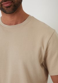 T-shirt beige a maniche corte con design a coste texturizzate, dotato di scollo rotondo e cuciture visibili lungo i bordi.