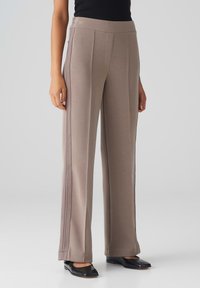 Wijdvallende broek in een zachte taupe stof, met een gladde textuur, zijdelingse pipingdetails en een aansluitende tailleband. Gepaarde met zwarte flats.